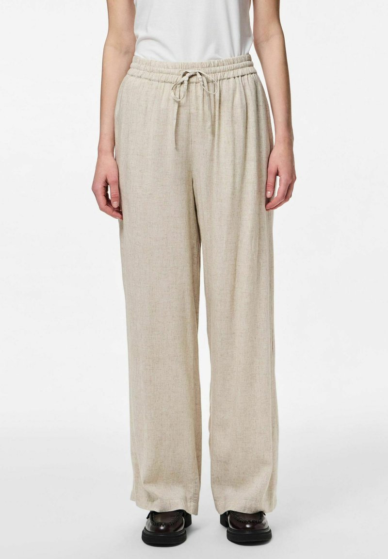 Pantalons beige à jambes larges avec une taille élastique et un cordon de serrage. Fabriqués en tissu léger avec une finition texturée. Associés à des chaussures noires.