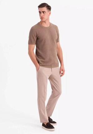 Suéter de punto marrón de mangas cortas, pantalones beige claros ajustados y zapatos oscuros sin cordones. El conjunto presenta un patrón de punto texturizado.