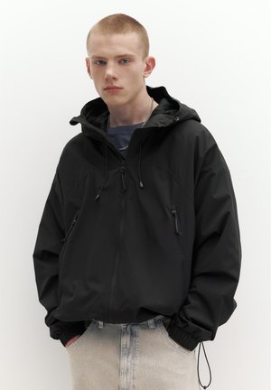 Mladý muž s krátkymi vlasmi v čiernej bunde s kapucňou a svetlomodrých džínsach, stojaci s rukami v vreckách na bielom pozadí.