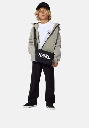Niño rubio de pie que lleva una gorra negra, chaqueta gris claro, camiseta blanca, pantalones negros, zapatillas negras y un bolso cruzado negro con la etiqueta "KARL".