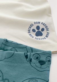 Paw Patrol-themed outfit featuring a pale cream top with a blue paw print logo and teal bottoms with printed character designs.