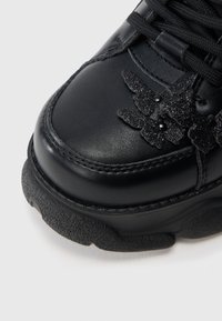 Gros plan sur une chaussure noire à semelle chunky texturée avec des appliques papillon scintillantes près des lacets noirs.