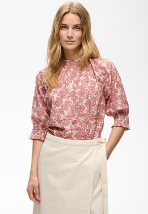 Femme aux longs cheveux blonds porte un chemisier fleuri rose à manches bouffantes et une jupe portefeuille crème, debout devant un fond blanc uni.