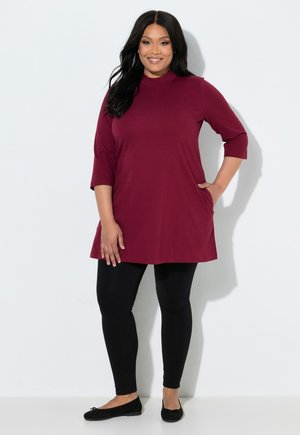 Kvinde smiler iført en burgunderrød tunika med lommer, sorte leggings og sorte flade sko, stående mod en hvid baggrund.