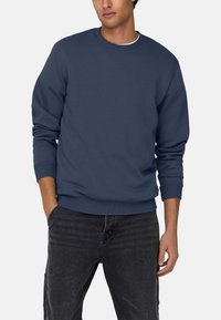 Sweat-shirt marine en mélange de coton, design à col rond, poignets et ourlet côtelés, texture lisse, porté avec un pantalon noir, mains dans les poches.