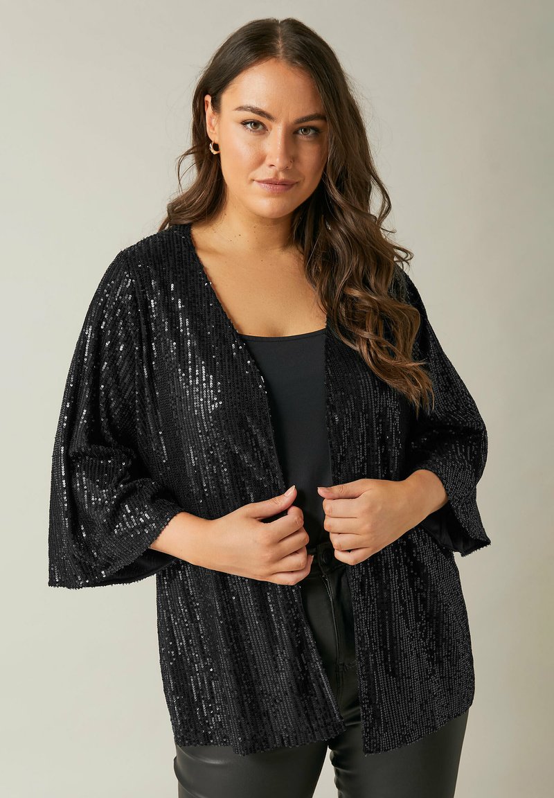 Kimono noir à sequins avec un devant ouvert, des manches larges et un motif à rayures verticales. Porté sur un top noir, avec une texture brillante.