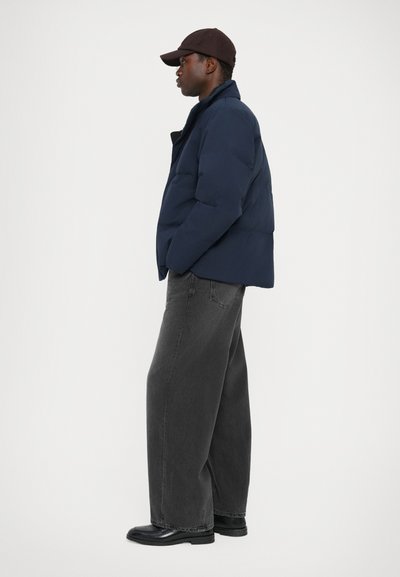 Navy puffer jacket with a high collar, side pockets, and a smooth texture, paired with loose-fitting dark gray jeans and black shoes.