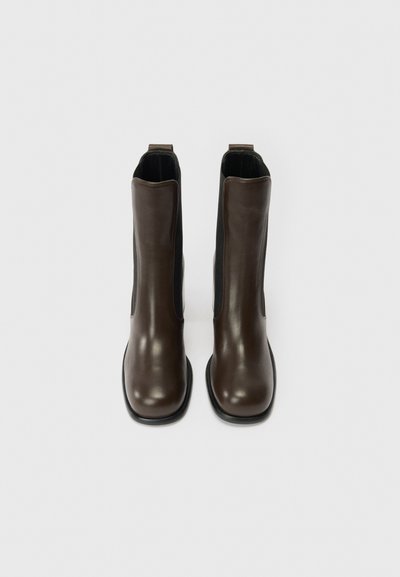 Σκούρες καφέ δερμάτινες μπότες μέχρι τον αστράγαλο με μαύρες ελαστικές πλευρικές πάνελ και λαβές, εμφανιζόμενες από το μπροστά σε λευκό φόντο.