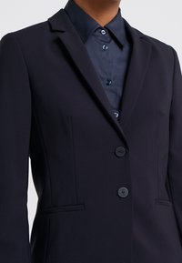 Blazer bleu marine en tissu lisse, doté d'un revers cranté, de trois boutons et de poches latérales ; porté sur une chemise sombre à boutons.