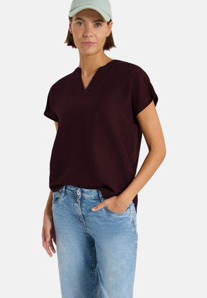 Femme portant une casquette vert clair, un haut ample à manches courtes violet foncé et un jean bleu clair, une main dans la poche, fond neutre.