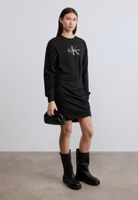 Μαύρο φόρεμα φούτερ με λογότυπο Calvin Klein, υφή υφάσματος, μακριά μανίκια, συνδυασμένο με μαύρες μπότες μέχρι το γόνατο και μικρή μαύρη τσάντα.