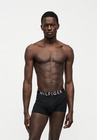 Homme musclé à la peau foncée portant un boxer noir Tommy Hilfiger, debout devant un fond clair uni avec une expression sérieuse.