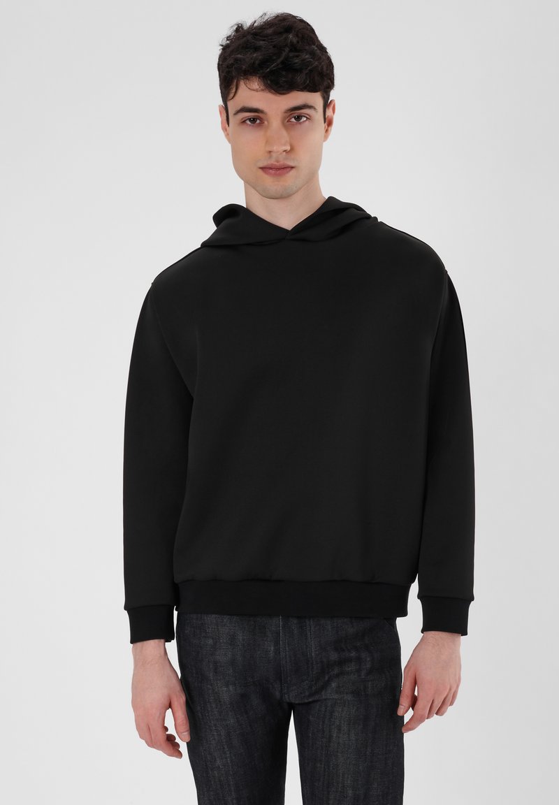 Schwarzer Hoodie mit lockerem Schnitt, gerippten cuffs und Taillenbund, aus glattem Stoff. Kombiniert mit dunklen Jeans.