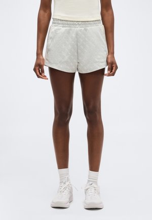 Inimene, kellel on seljas heledat halli mustriga spordipüksid, valge tanktop, valged sokid ja valged tossud, seisab ühevärvilise tausta ees.