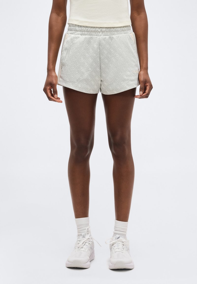 Άτομο που φοράει αθλητικές σορτς σε ανοιχτό γκρι με σχέδια, λευκή φανέλα, λευκές κάλτσες και λευκά αθλητικά παπούτσια, στέκεται μπροστά σε ένα απλό φόντο.