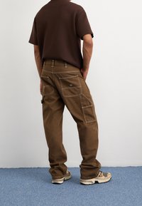 Pantalon cargo marron en tissu robuste, doté de multiples poches et de coutures contrastantes. Porté avec une chemise marron à manches courtes et des baskets.