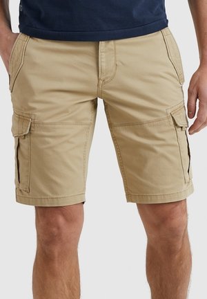 Uomo che indossa pantaloncini cargo beige con tasche laterali con patta e una camicia blu scuro, in piedi con le mani vicino alle tasche.
