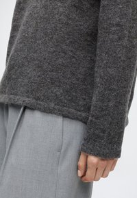Personne portant un pull gris foncé en matière duveteuse et un pantalon gris clair, avec la main reposant sur le côté.