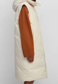 Gilet crème matelassé avec capuche et fermeture éclair, comportant des manches en pull tricoté beige et des poches latérales. Texture lisse et design allant jusqu'aux genoux.