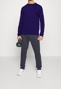 Man i mörklila långärmad skjorta, mörkgrå Adidas sweatpants, vita sneakers, håller en svart kettlebell med en blå rand.