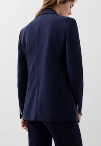 Navy blazer s přizpůsobeným střihem, klopovým límcem a nenápadným polstrováním na ramenou. Disponuje větraným zadním dílem a vzorovanými manžetami vykukujícími z rukávů.