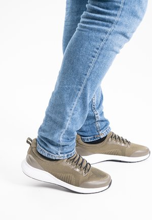 Person trägt hellblaue Jeans und beige Sportschuhe mit weißen Sohlen und steht mit verschränkten Beinen vor weißem Hintergrund.