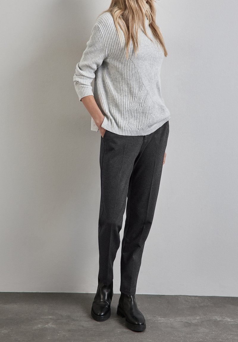 Personne portant un pull côtelé gris clair, un pantalon fuselé gris foncé et des bottines noires, debout contre un mur clair.