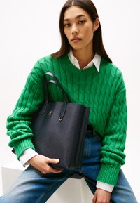 Sort struktureret tote taske med to hanke, der har et subtilt mønster og guld hardware, holdt af en person i en grøn kabelstrikket sweater.