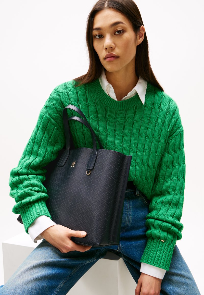 Sort struktureret tote taske med to hanke, der har et subtilt mønster og guld hardware, holdt af en person i en grøn kabelstrikket sweater.