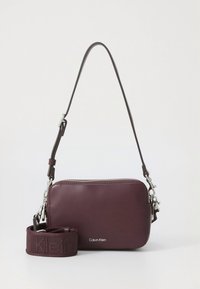 Sac bandoulière en cuir bordeaux profond avec une fermeture éclair et des détails métalliques argentés. Comprend une bandoulière en tissu amovible avec logo.