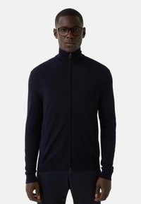 Mężczyzna w czarnych oprawkach okularów i granatowym swetrze na zamek, stojący na tle białym, zwrócony przodem z neutralnym wyrazem twarzy.