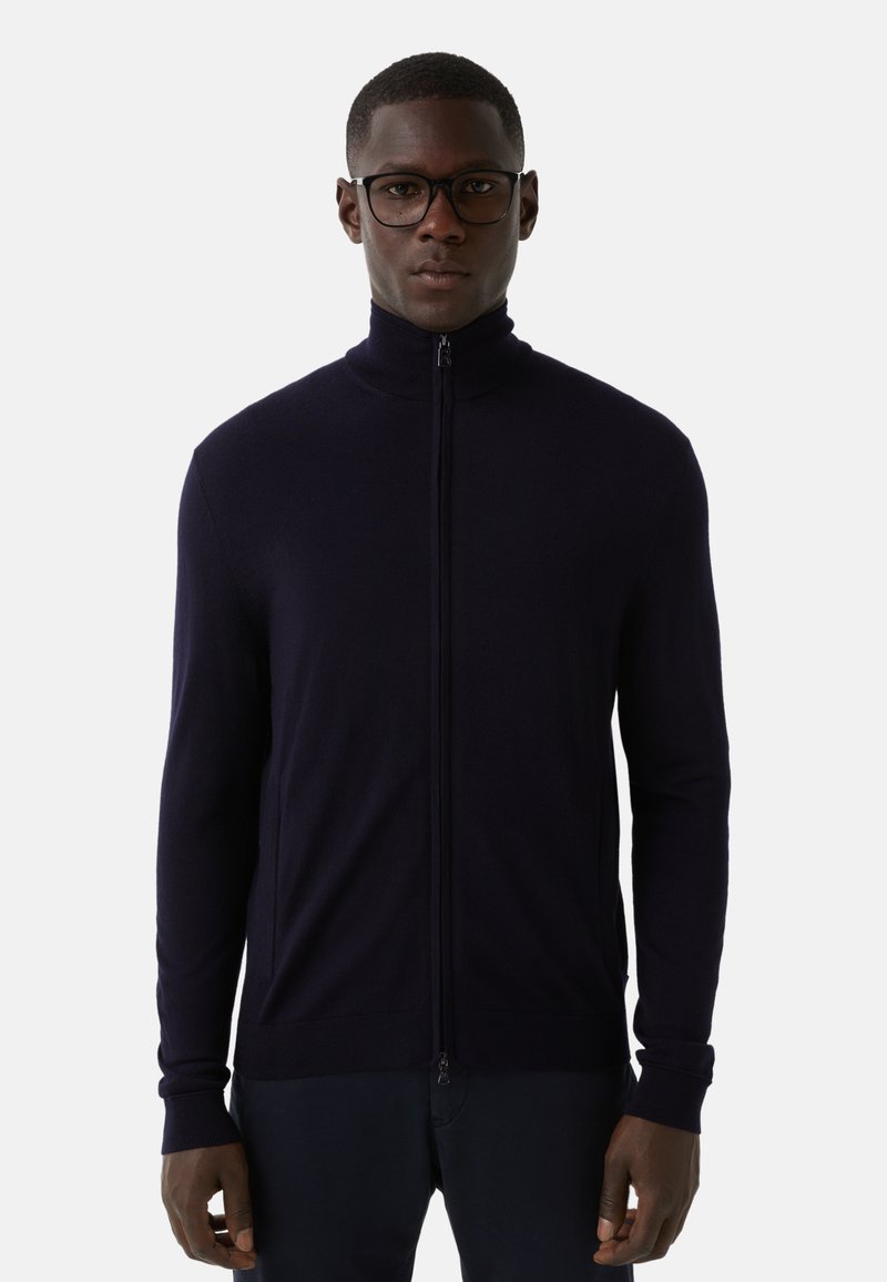 Mężczyzna w czarnych oprawkach okularów i granatowym swetrze na zamek, stojący na tle białym, zwrócony przodem z neutralnym wyrazem twarzy.