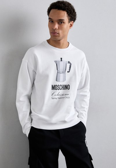 Sudadera blanca con un gráfico de una cafetera moka y el texto "MOSCHINO Colazione Primavera Verano 2025", combinada con pantalones negros.