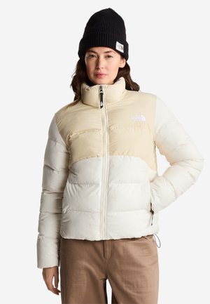 Frau trägt eine beige-weiße Steppjacke, schwarze Strickmütze und braune Hose, steht mit einer Hand in der Tasche auf weißem Hintergrund.
