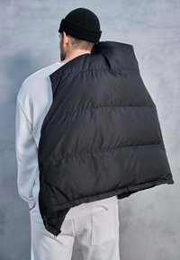Doudoune noire oversize avec un col haut, présentant des sections matelassées et un design asymétrique, portée sur un léger pull et un pantalon.