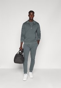 Felpa grigia con zip e pantaloni jogger coordinati, realizzati in tessuto morbido. Sneakers bianche e una borsa a tracolla nera completano il look.