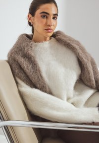 Pull en tricot moelleux crème et marron superposé à un châle marron assorti. Le tissu semble doux avec une texture à mailles lâches. Une chaise métallique est visible.