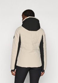Casaco puffer em bege e preto com gola alta, apresentando um design acolchoado, painéis laterais e um logotipo visível na manga.