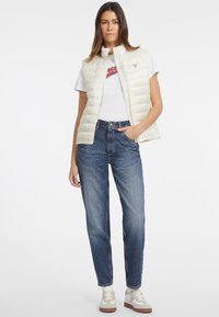 Helle cremefarbene Steppweste mit hohem Kragen, getragen über einem weißen T-Shirt mit rotem Grafikdruck. Kombiniert mit lockeren blauen Jeans und beigen Sneakers.