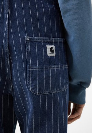 Spodnie dżinsowe z białymi pionowymi prążkami, kwadratową kieszenią z naszywką z logo Carhartt oraz osobą w niebieskiej koszuli z długim rękawem.