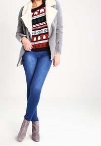 Grå ullblandad kappa med en shearlingkrage, kombinerad med en svart jultröja med färgglada mönster, smala blå jeans och grå ankelstövlar.