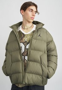 Young man wearing an olive green puffer jacket over a graphic sports jersey, looking sideways, with silver chain necklace and earring.