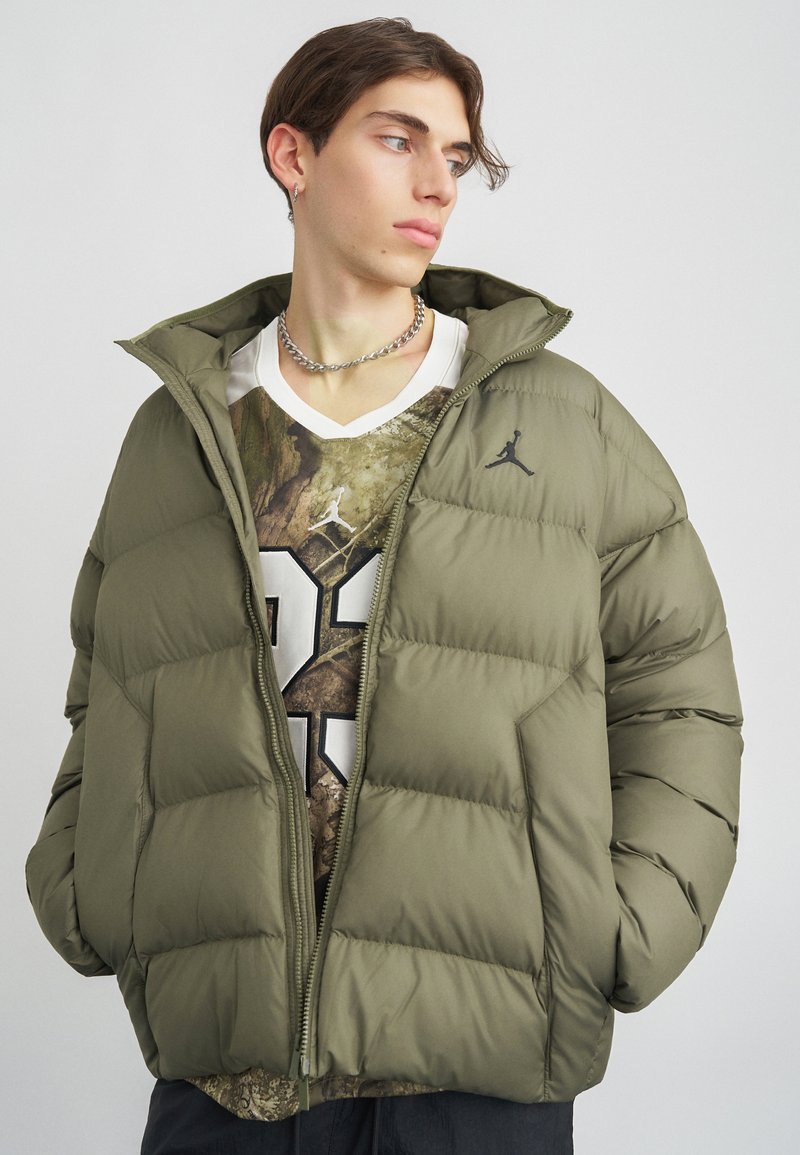 Young man wearing an olive green puffer jacket over a graphic sports jersey, looking sideways, with silver chain necklace and earring.
