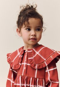 Vestito scozzese rosso con colletto arricciato, chiusura con bottoni e tessuto strutturato. Progettato con strisce rosa e bianche che formano il motivo a quadri.