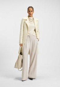 Hellbeige Wollmantel über einem cremefarbenen Pullover, kombiniert mit weit geschnittenen beigen Hosen und einer passenden Handtasche. Spitz zulaufende schwarze Absätze runden den Look ab.