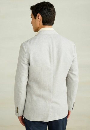 Bărbat văzut din spate purtând un sacou texturat gri deschis cu gulere de cămașă bej și pantaloni închisi la culoare, pe un fundal simplu și deschis la culoare.