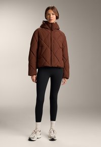 Brun quiltet pufferjakke med hætte, der har et diamantmønster og frontlynlås, parret med sorte leggings og sneakers.