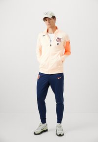 Light peach zip-up jacket with orange sleeve, featuring a logo. Dark blue joggers with orange accent. White and gray athletic shoes.