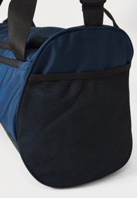 Blå duffelbag med en teksturert mørk mesh-bunn og svarte stropper, med en avrundet form og glatt, slitesterk stoff.