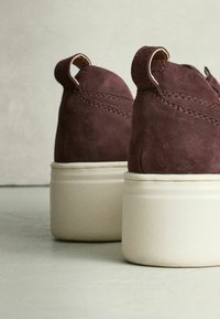 Burgundy Wildleder-Sneaker mit abgerundeter Form, ausgestattet mit einer weißen, strukturierten Plateausohle und kleinen Zugschlaufen an der Ferse.